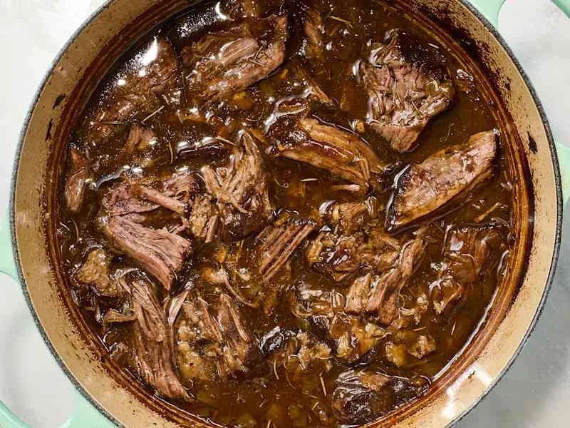 Tender shredded beef in a rich brown sauce, simmering in a large pot with herbs.