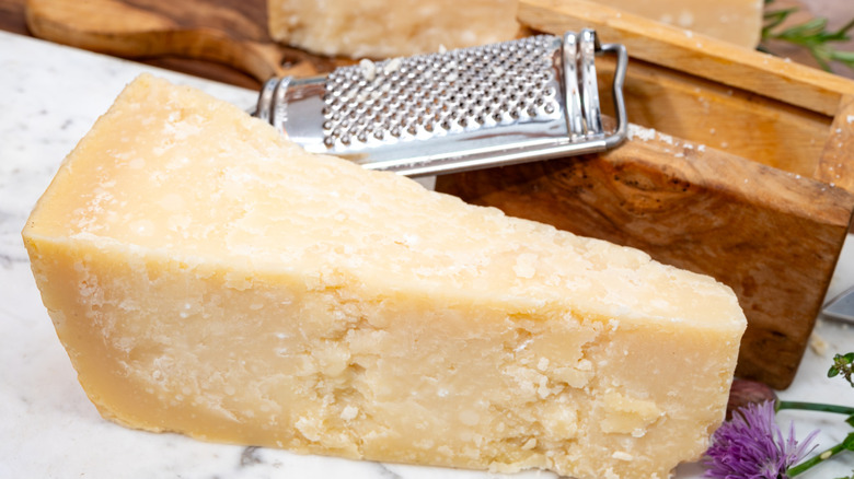 A wedge of Parmigiano Reggiano cheese next to a wooden box with a purple flower in the foreground and a cheese grater in the background