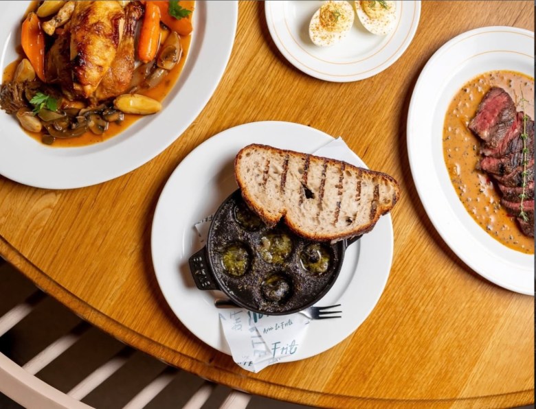 Overhead view of French bistro dishes on a wooden table: escargot with grilled bread, roasted chicken, steak au poivre, and deviled egg.