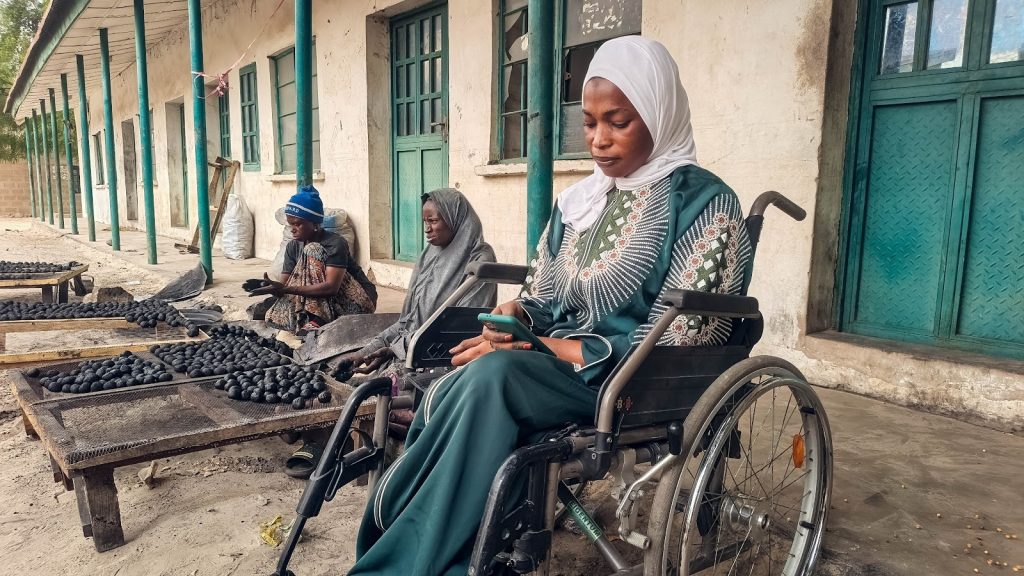 Three women are outside a building. One in a wheelchair uses a phone, while two others sort charcoal balls.