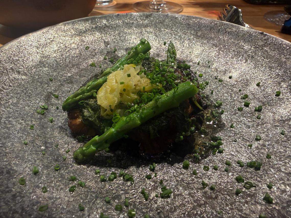 A close-up of a seared mushroom fillet served on a dark, textured stone plate. It is topped with a generous layer of green herb chimichurri, bright green asparagus spears, and caramelized onions, all garnished with fresh chives.