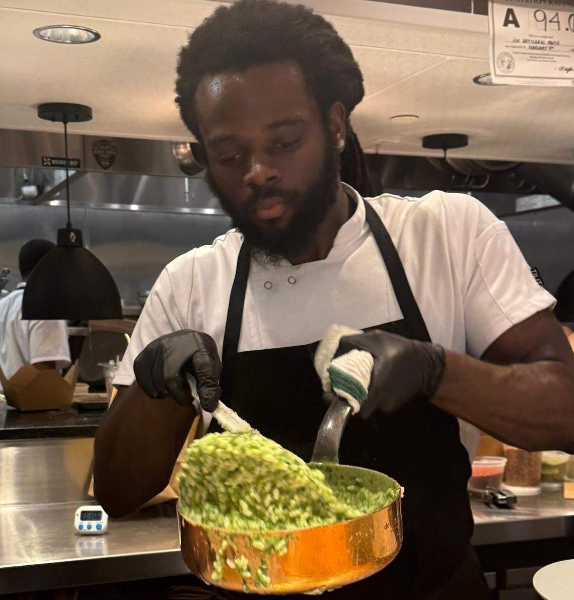 A chef with long dreadlocks and a white uniform vigorously stirring a copper pot filled with bright green risotto. He is wearing black gloves and working in a professional kitchen setting with a health inspection “A” rating visible in the corner.
