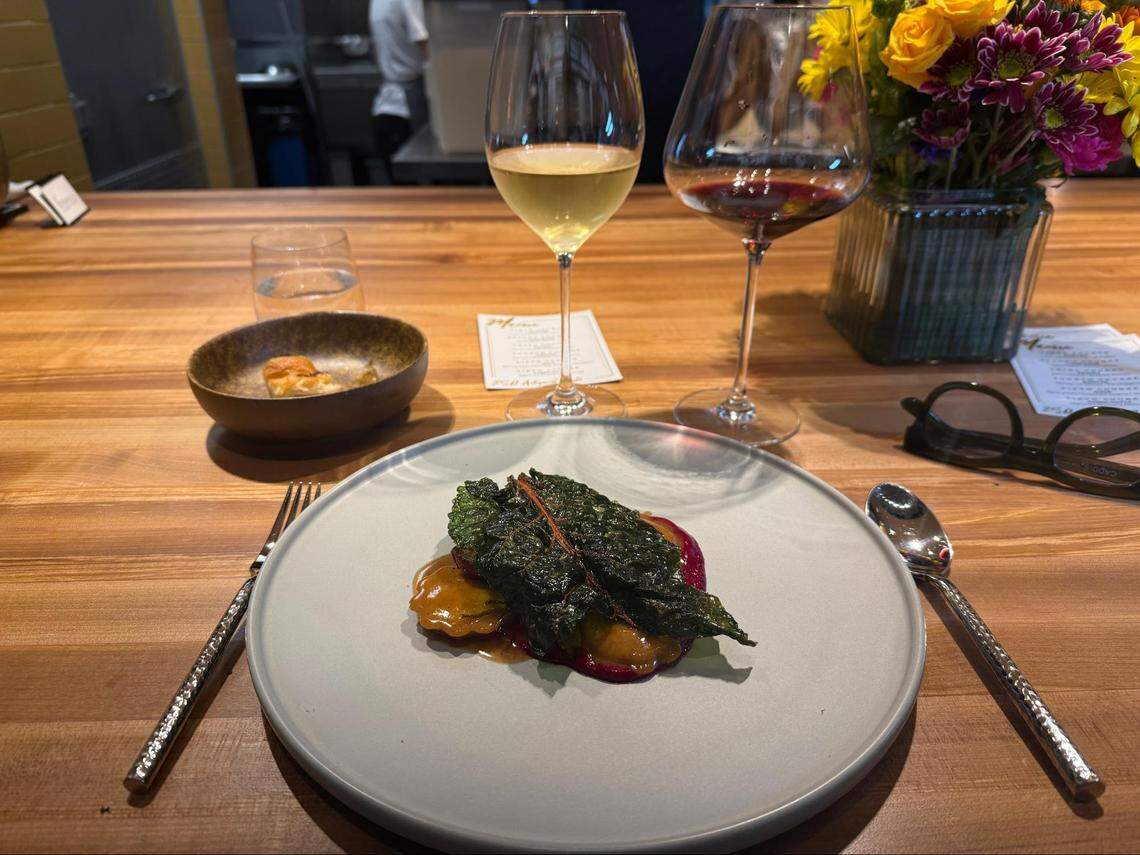 An elegantly plated ravioli dish topped with a large, dark green wilted kale leaf and a rich brown sauce. The dish is served on a gray plate at a wooden bar, flanked by a glass of white wine and a glass of red wine.