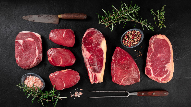Various cuts of beef beside knives, spices, and sprigs of herbs