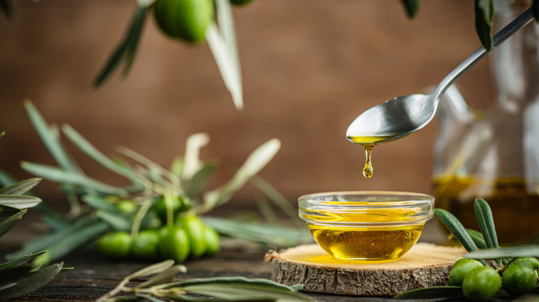 A small bowl of olive oil with olive branches scattered around and a spoon dripping oil back into the bowl