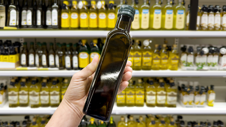 Someone holding up a glass bottle of olive oil in front of grocery store shelves filled with olive oil