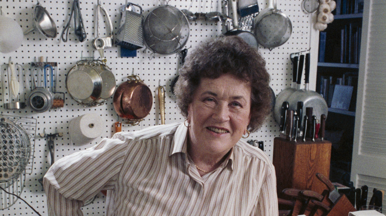 Julia Child in kitchen with kitchen utensils hanging on a wall behind her
