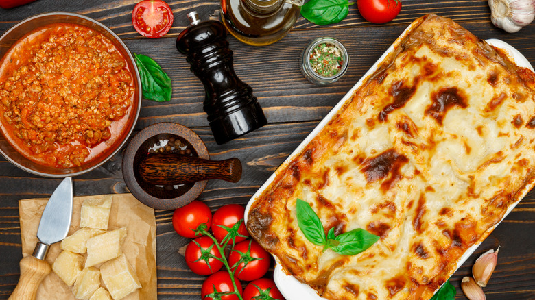 whole homemade lasagne with ingredients around it, including tomatoes, dried herbs, pepper, meat sauce, and parmesan