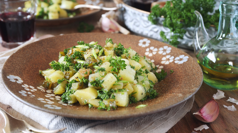 Rustic French potato salad with fresh herbs on a brown plate next to ingredients and wine glass