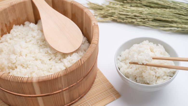 Cooked sushi rice in a large wooden tub with a wooden spoon and in a smaller white bowl with chopsticks