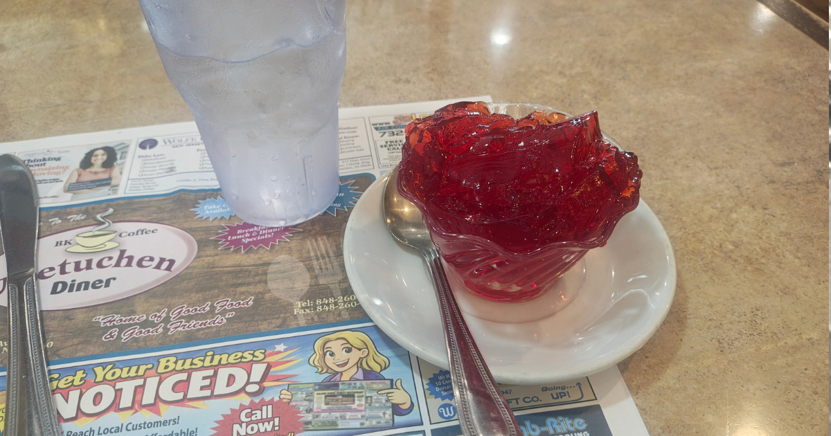 A small glass cup of red jello on a plate, next to a glass of Sprite. 