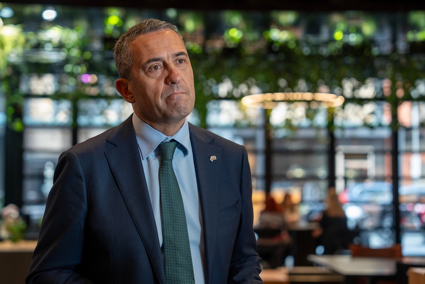 Tom Koutsantonis pursed lips in a serious expression in a room with large glass windows and plants hung from the ceiling