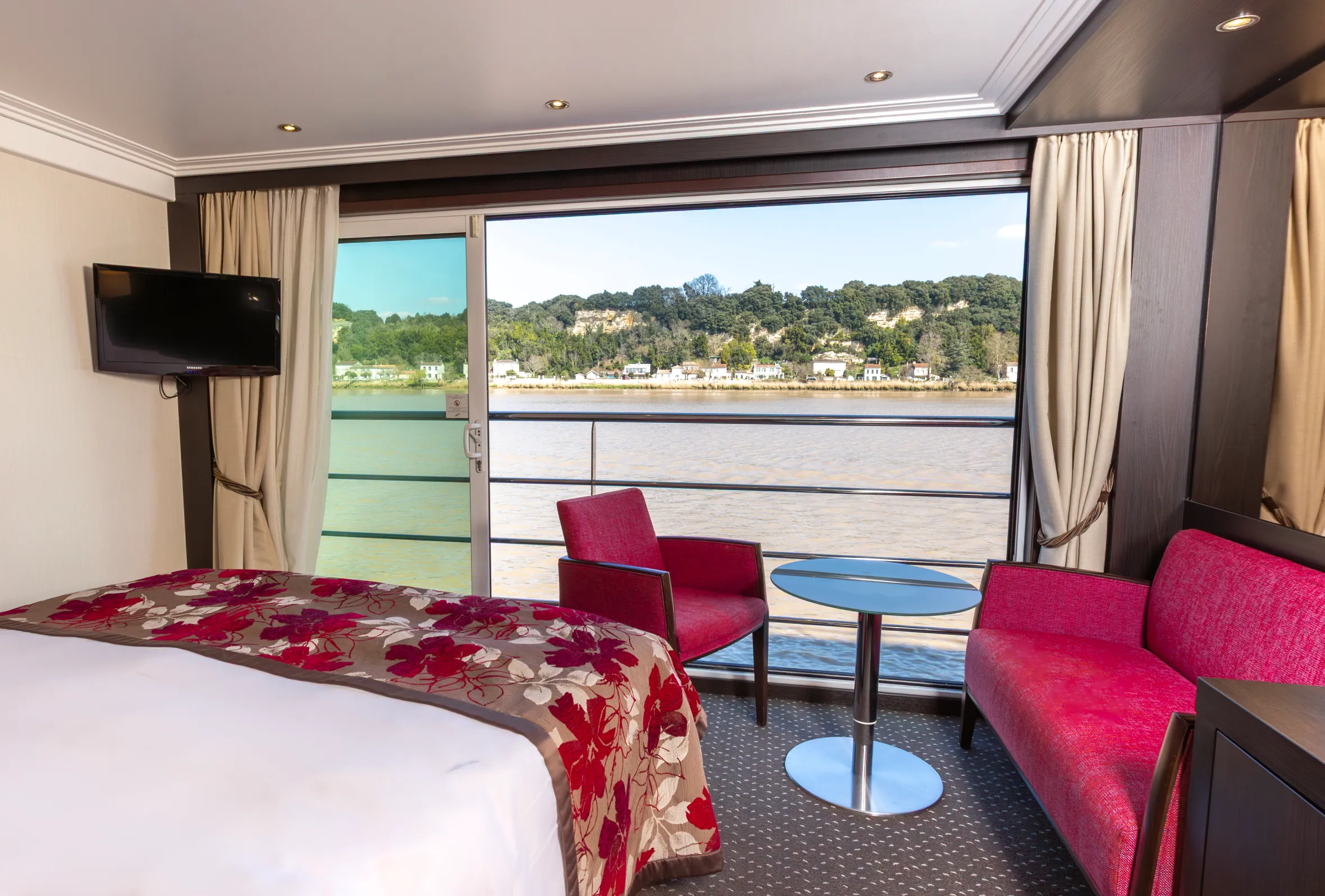 Cabin aboard the Avalon Artistry II river cruise ship with a bed, red seating, and large windows overlooking a river and treed landscape.
