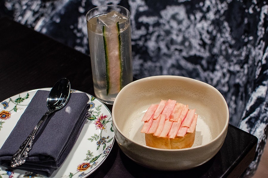 A dessert with thin pink slices arranged on top, served in a beige bowl. Next to it is a floral-patterned plate with a folded dark napkin and a silver spoon. Behind the bowl is a tall glass of clear liquid with ice and two long cucumber slices.