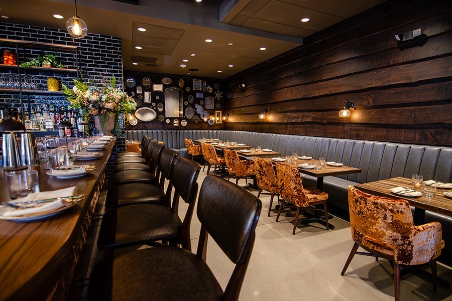 A modern restaurant interior featuring a long wooden bar with black leather bar stools on one side and a row of tables with plush, burnt orange velvet chairs on the other. The back wall is covered with dark wood paneling and a long, tufted gray banquette. The opposite wall has black brick with shelves holding bottles and decorative items, and a large floral arrangement in a vase on the bar. The space is warmly lit with hanging pendant lights and wall sconces.