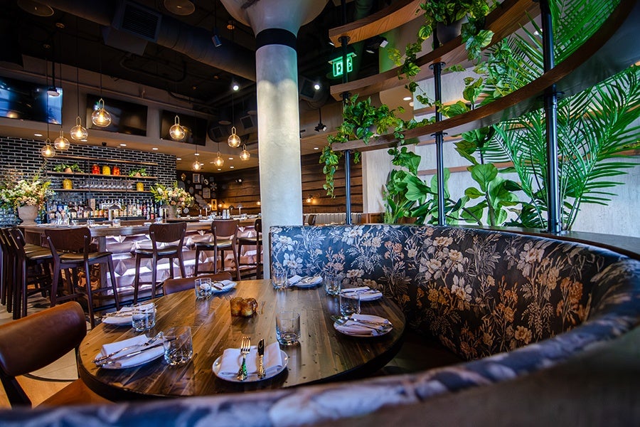 A cozy restaurant interior featuring a round wooden table set with plates, glasses, and silverware. Surrounding the table is a curved bench upholstered with floral-patterned fabric. Behind the seating area, there are green plants on a wooden shelf. In the background, a bar with high chairs, hanging pendant lights, and shelves stocked with bottles and glasses is visible. The ceiling has exposed ducts and spotlights.