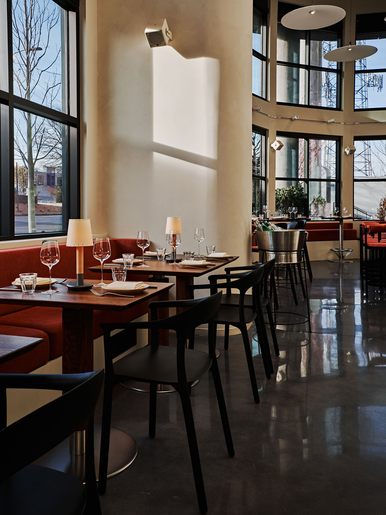 Modern restaurant interior with set tables, black chairs, red cushioned seating, large windows, and natural light reflecting on polished floors.