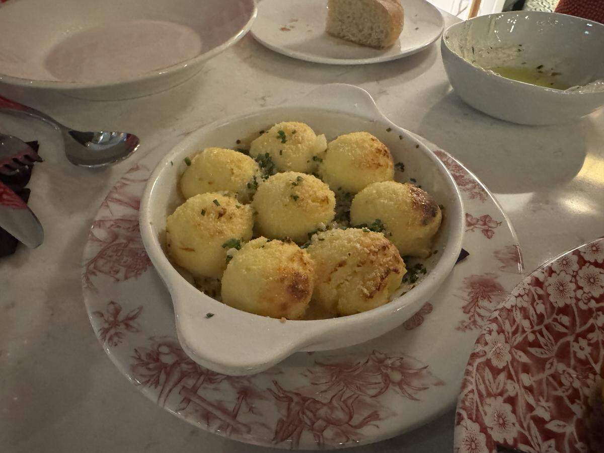 A small, white ceramic baking dish with handles, containing about eight round, golden-brown gnocchi or baked semolina dumplings. The dumplings are lightly browned on top, dusted with cheese, and sprinkled with finely chopped chives. The dish rests on a white plate with a red floral design.