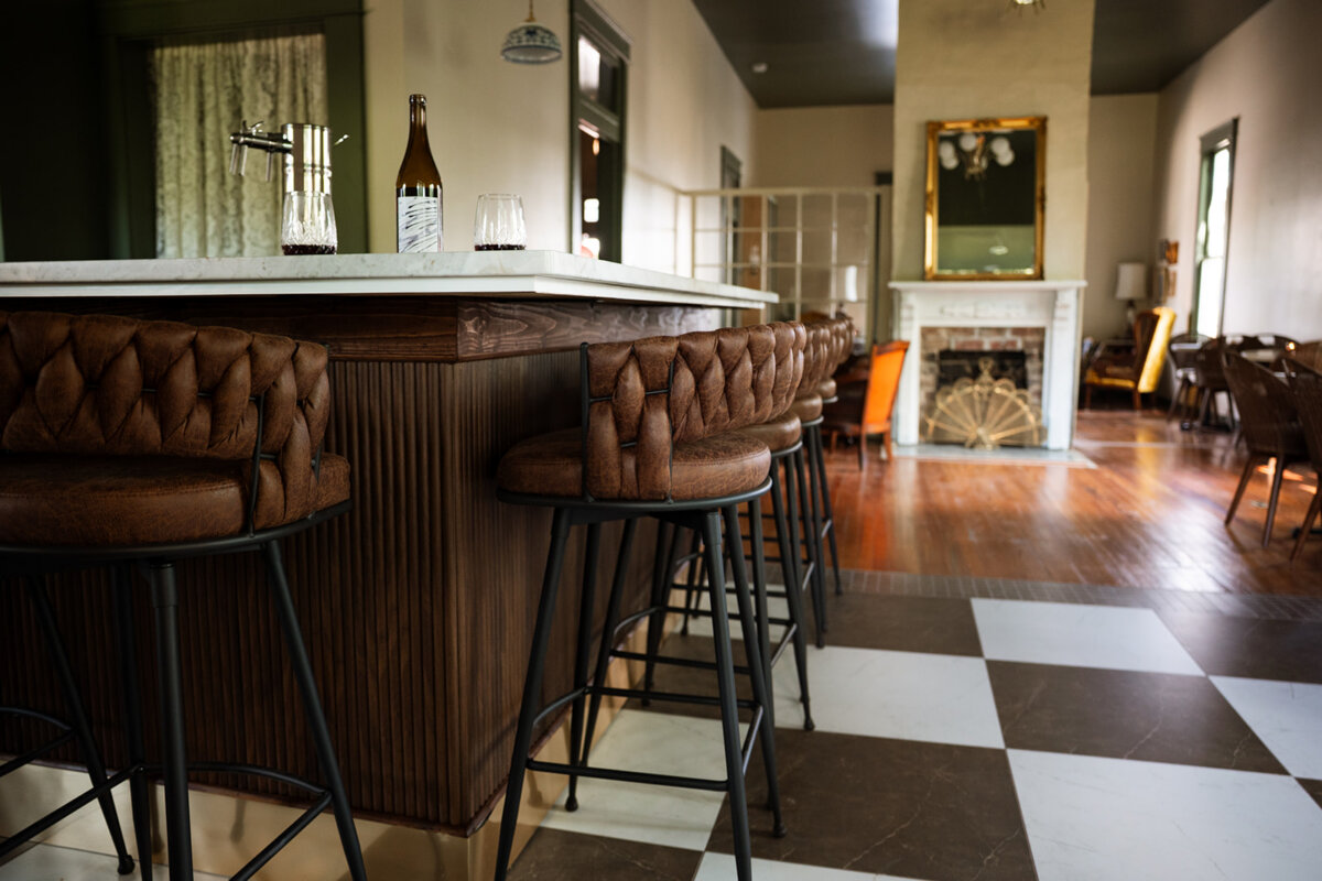 A bar area with tufted leather stools, a marble countertop, a bottle and glasses, and a view into a spacious Lauren Maranto-designed lounge with a fireplace and wooden floors.