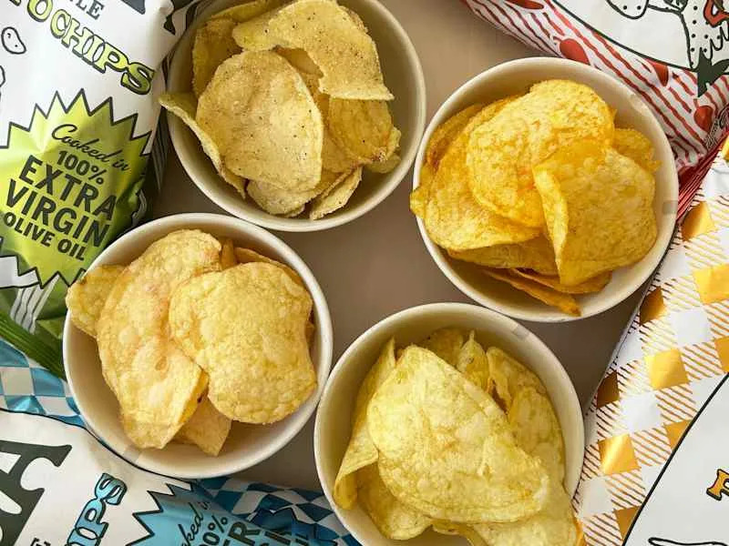 Assorted potato chips in bowls, featuring different textures and colors, with colorful packaging in the background.