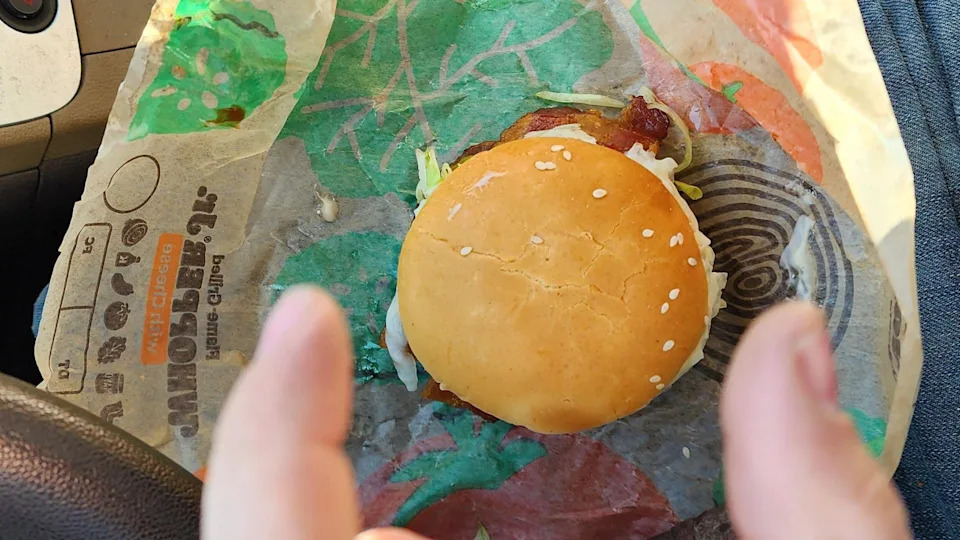 A sesame seed hamburger on fast food wrapper sits on someone's lap in a car, with two fingers visible in the foreground about to grab the burger.