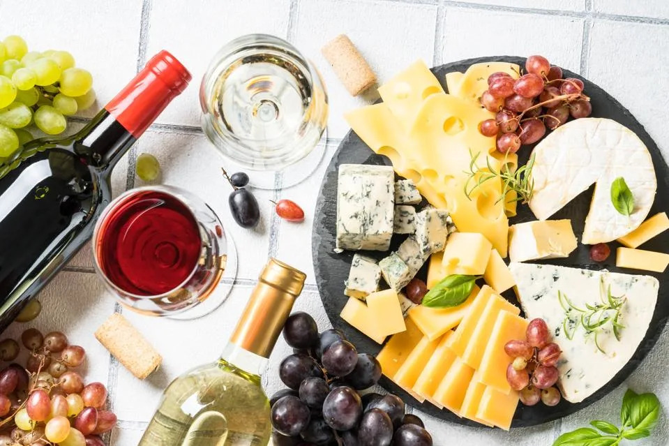 Cheese platter with craft cheese assortment and wine glasses at white tile background.