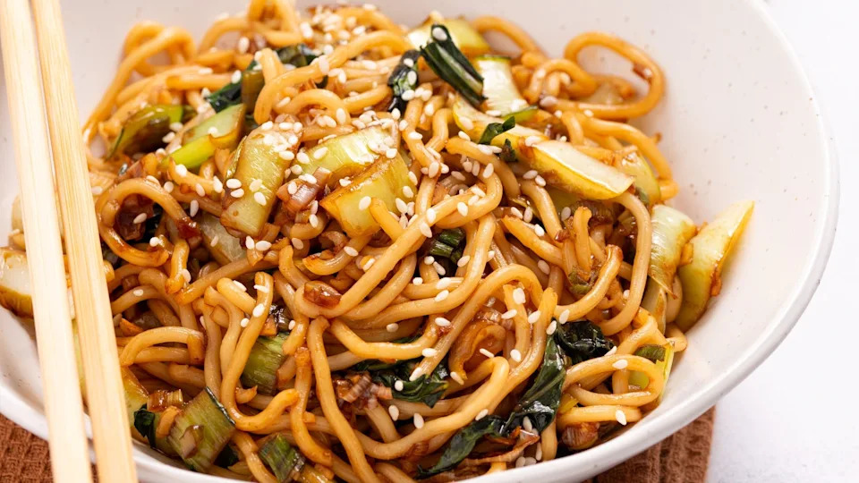 A bowl of stir-fried noodles with vegetables, topped with sesame seeds.  Chopsticks rest on the bowl's edge.
