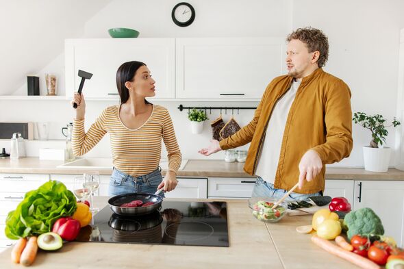Young couple having conflict while cooking together in their kitchen, woman swinging kitchen spatula at her husband, man preparing salad