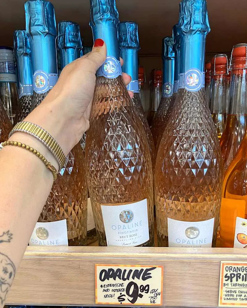 A hand holding a bottle of Opaline Brut Rosé wine with a blue foil cap, displayed on a shelf with other bottles.