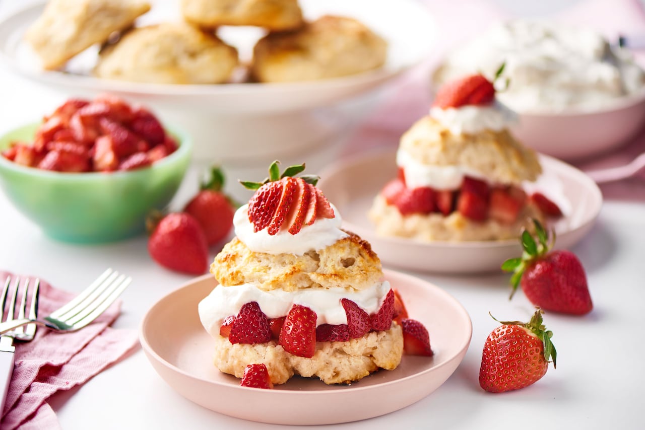 Sliced strawberries and whipped cream in between a scone sliced in half, and topped with more whipped cream. The strawberry shortcake is sitting on a shallow pink bowl on a table with more scones and berries in the background. 