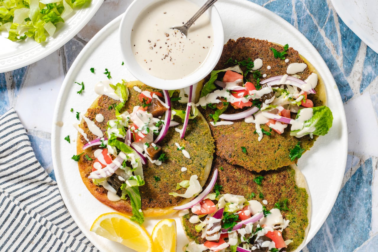 An overhead shot of a plate of falafel smash tacos and a small bowl of tahini sauce on a blue countertop.