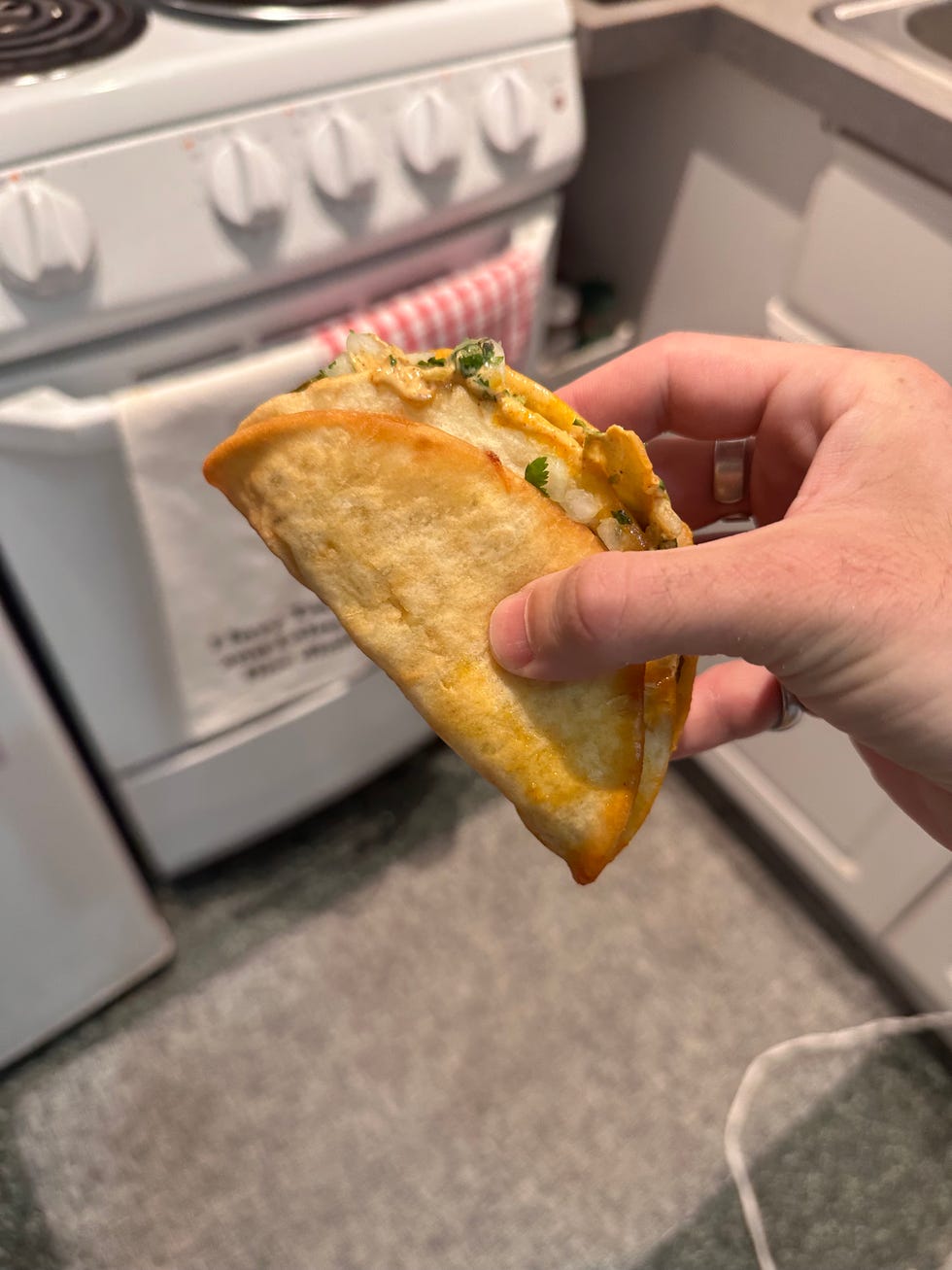 Hand holding a cheesy taco near a kitchen stove. Hand holding a cheesy taco near a kitchen stove.