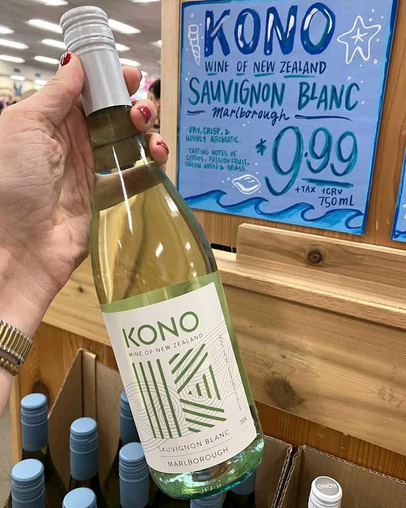 A hand holding a bottle of Kono Sauvignon Blanc wine with a green label, displayed near a promotional sign.