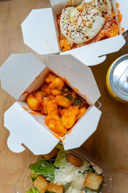 Creamy vodka sauce gnocchi served in a takeout container alongside a fresh Caesar salad with romaine lettuce, shaved Parmesan, and croutons at Gnocchi on 9th in Jersey City, New Jersey