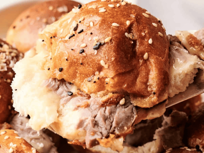 Close-up of a fork lifting a beef sandwich topped with sesame seeds on a soft bun, with more sandwiches in the background.