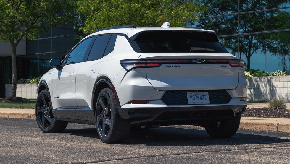 2026 Chevrolet Equinox EV 3RS rear rear three quarter view of a white suv parked in front of a modern glass building surrounded by trees.