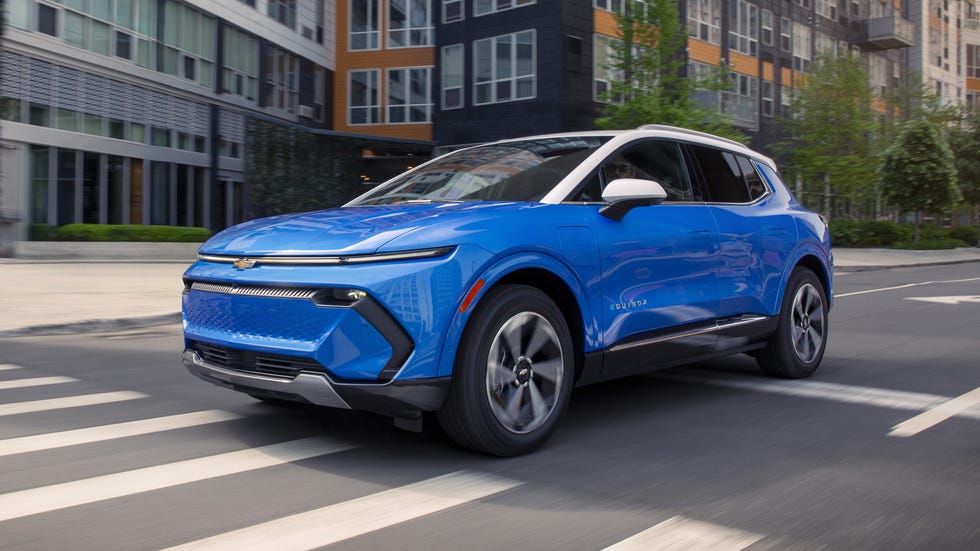 2027 Chevrolet Equinox EV front three quarter view blue suv driving through city street with modern apartment buildings in background.