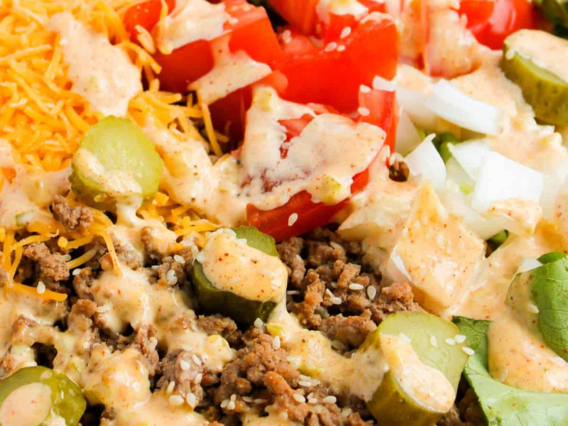 Close-up of a salad featuring ground beef, sliced pickles, diced tomatoes, shredded cheese, and diced onions, topped with a creamy dressing.