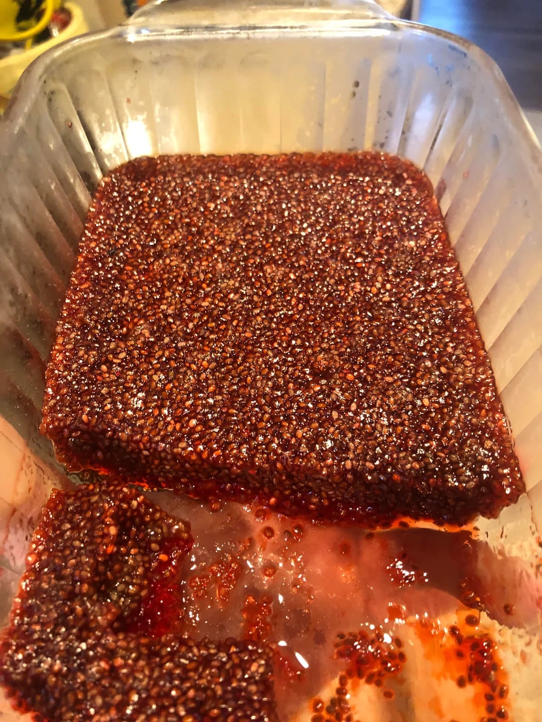 A close-up of a glass dish filled with a red, seed-filled gelatinous block, with a square piece cut out and sitting in juice at the bottom.