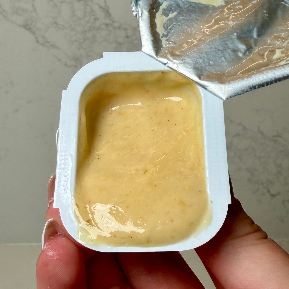 Open container of creamy sauce held in a hand, with a partially removed foil lid. Open container of creamy sauce held in a hand, with a partially removed foil lid.