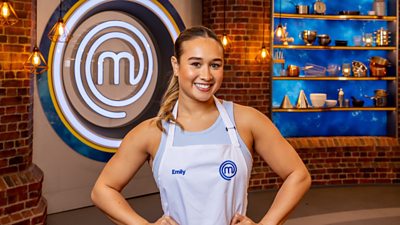 A woman wearing a light blue vest and a white apron stands in the masterchef studio smiling at the camera