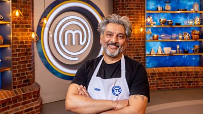 A man in a black t-short and white apron stands crossed arms smiling at the camera in the masterchef studio