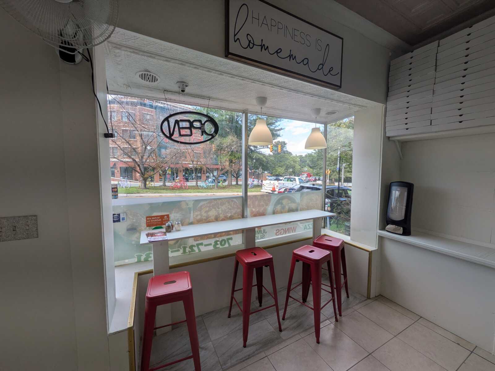 Interior of a small cafe counter with red metal stools and a white high table by a large window showing a street outside.