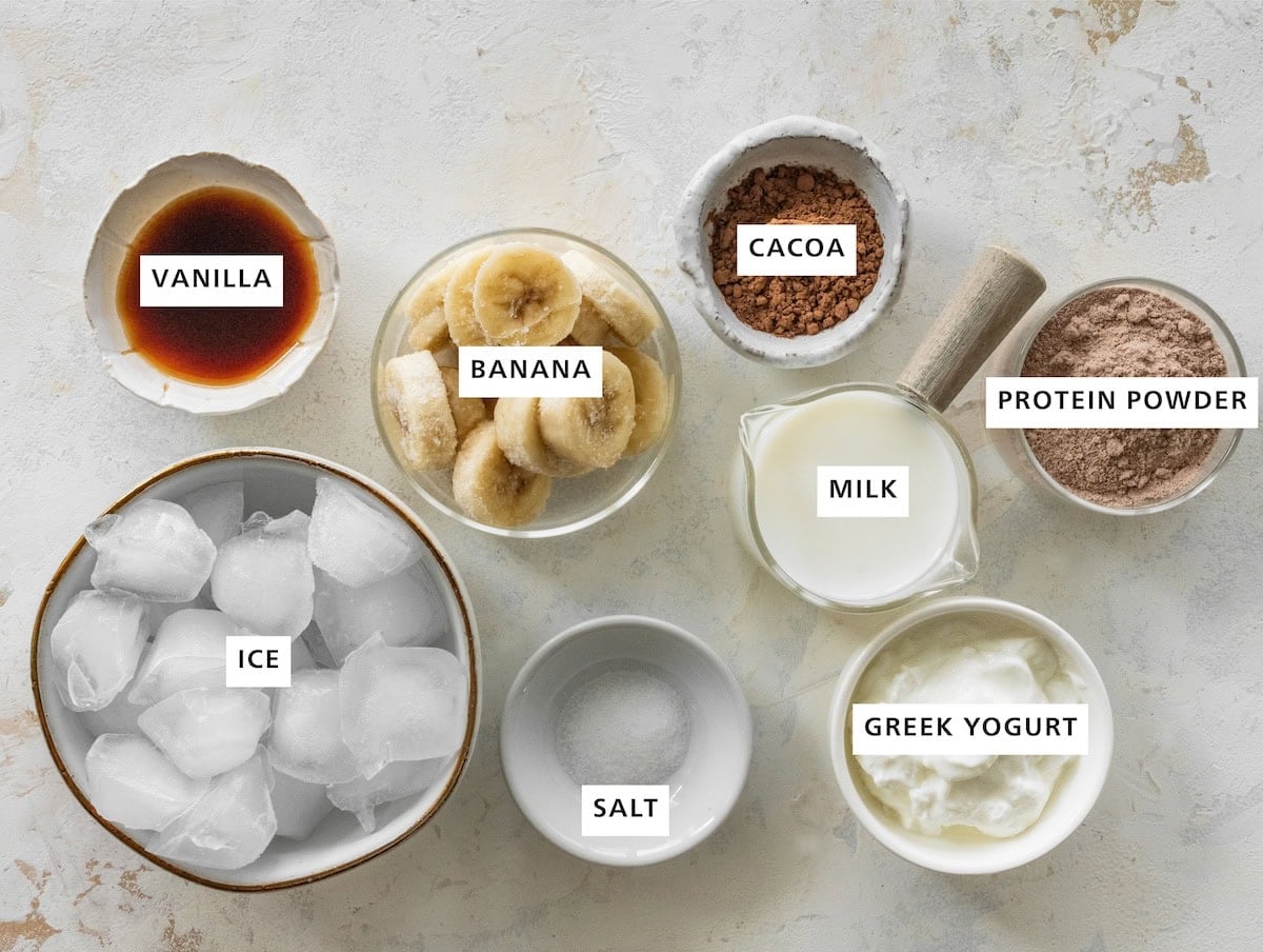 Ingredients measured out for a high protein copycat Wendy's frosty: Ice, salt, greek yogurt, milk, protein powder, cacoa, banana, and vanilla.