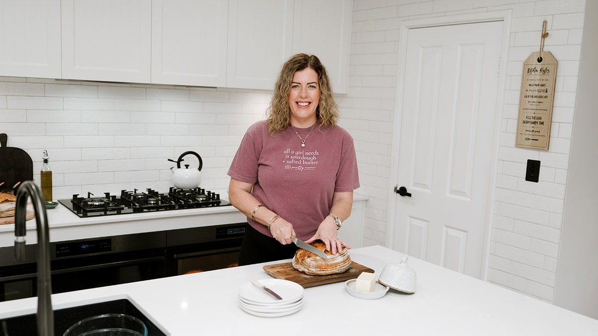 Kate Freebairn holds a knife over a piece of sourdough bread while wearing a shirt that reads, "All a girl needs is sourdough + salted butter."