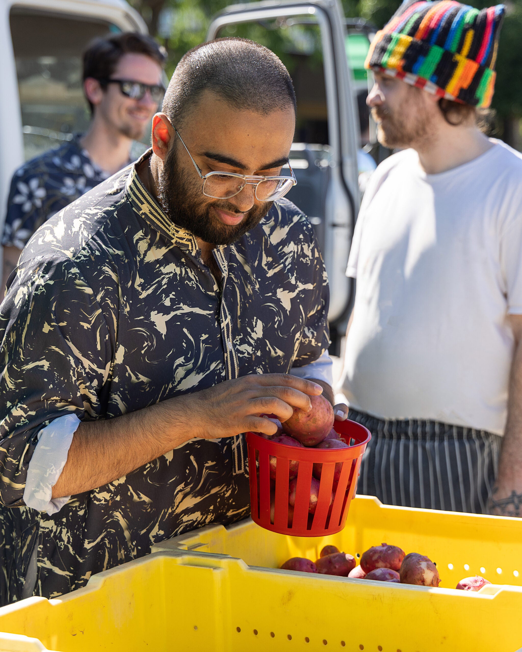 A person wearing glasses places apples into a red basket at an outdoor market, with two others standing in the background near yellow bins.
