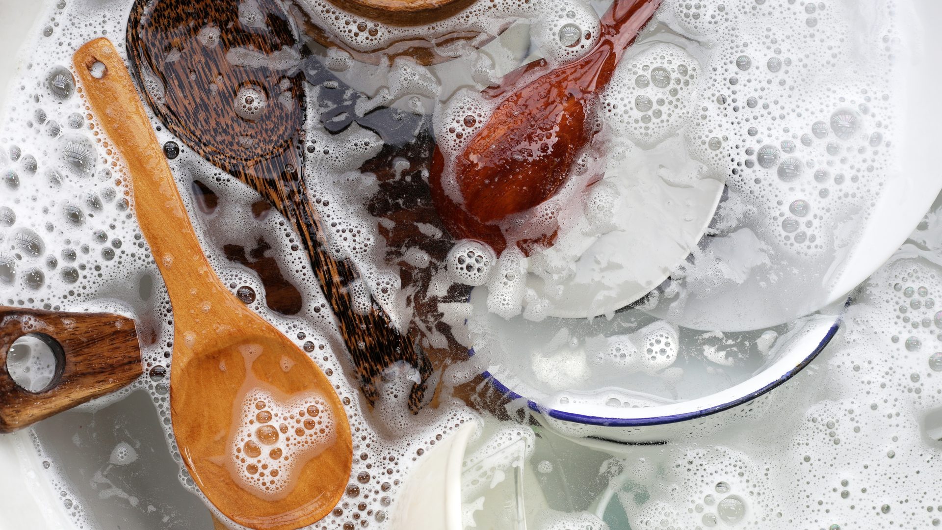 Overhead close-up of several wooden cooking spoons and utensils soaking in a white sink filled with soapy water and thick foam bubbles, alongside white and blue-rimmed enamel dishes being washed.