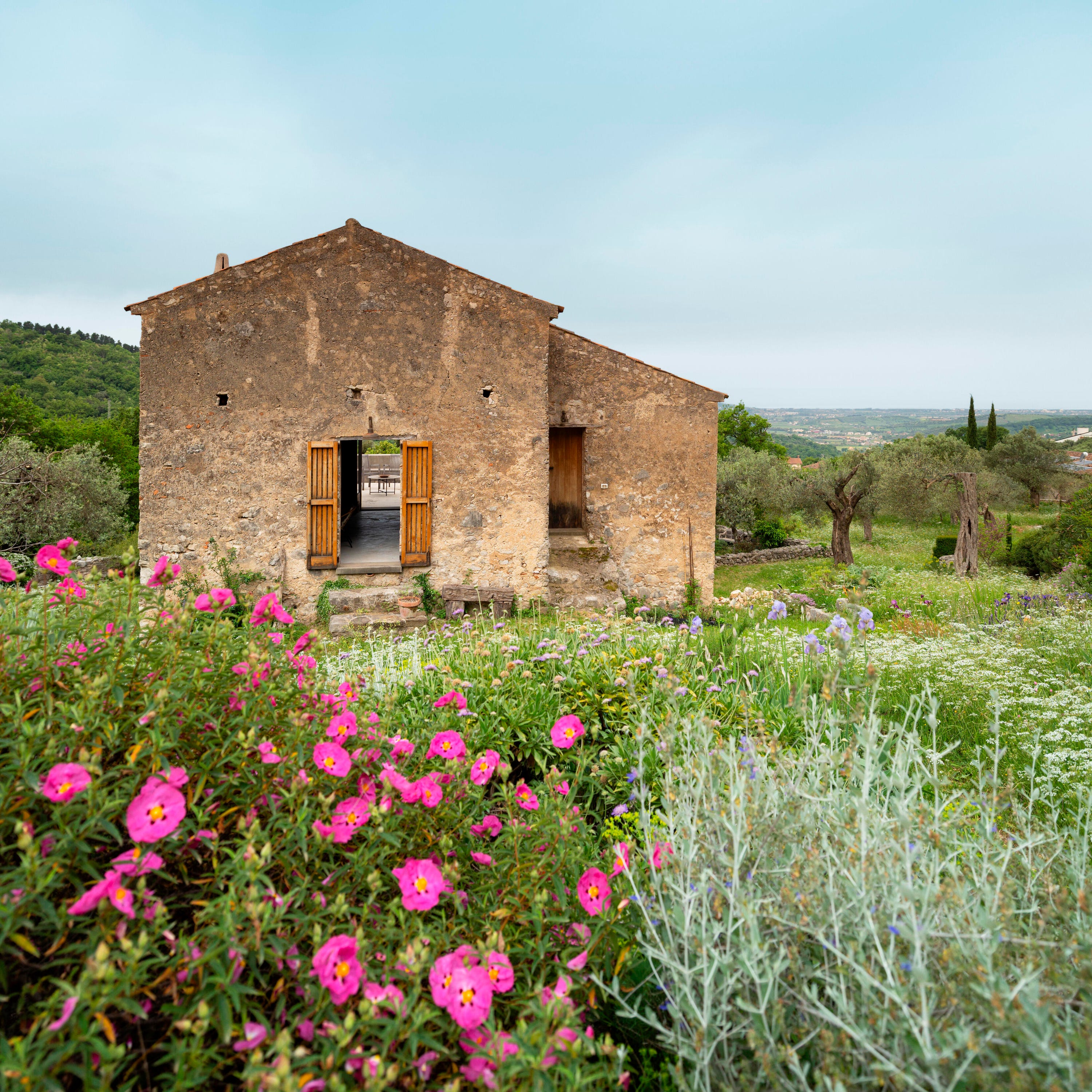 Rural stone house surrounded by vibrant flowers and greenery under a cloudy sky.